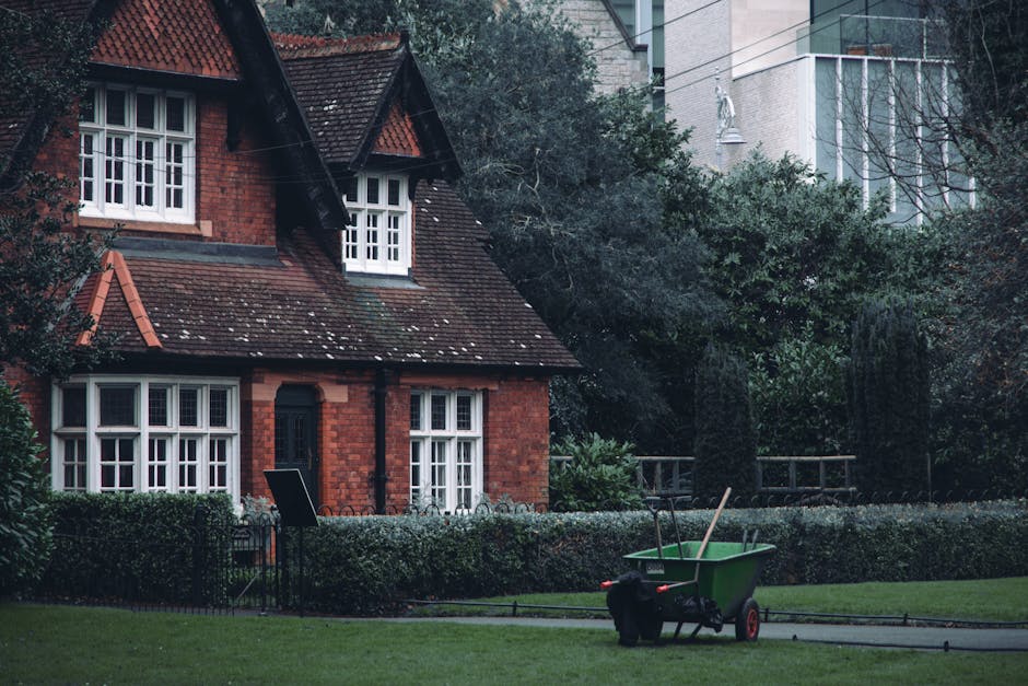 A front garden scene featuring a neatly maintained lawn with trimmed grass and a small border hedge surrounding the house. The house is a traditional red brick structure with white-framed windows and 