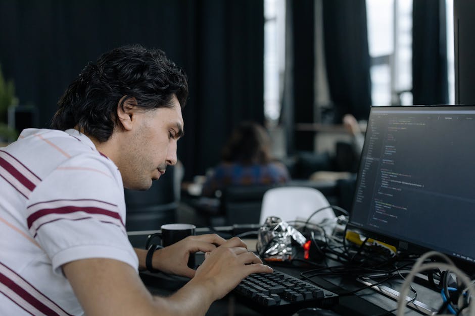 A young man with curly dark hair, wearing a white polo shirt with maroon and gray stripes, is intently working on a computer in an indoor workspace with large windows allowing natural light to brighte
