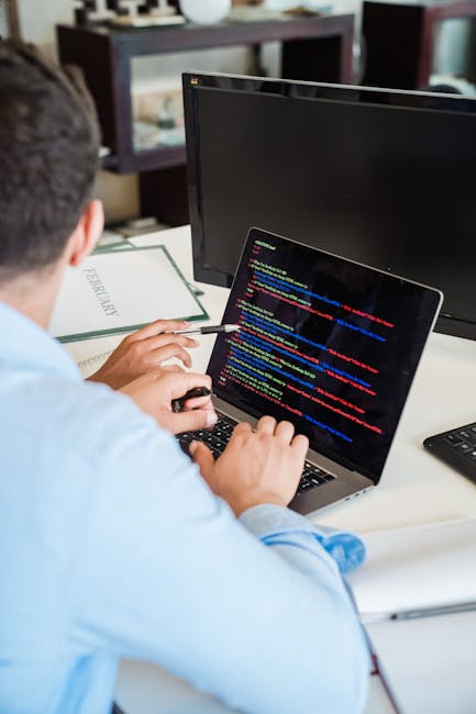 A young man with curly dark hair, wearing a white polo shirt with maroon and gray stripes, is intently working on a computer in an indoor workspace with large windows allowing natural light to brighte